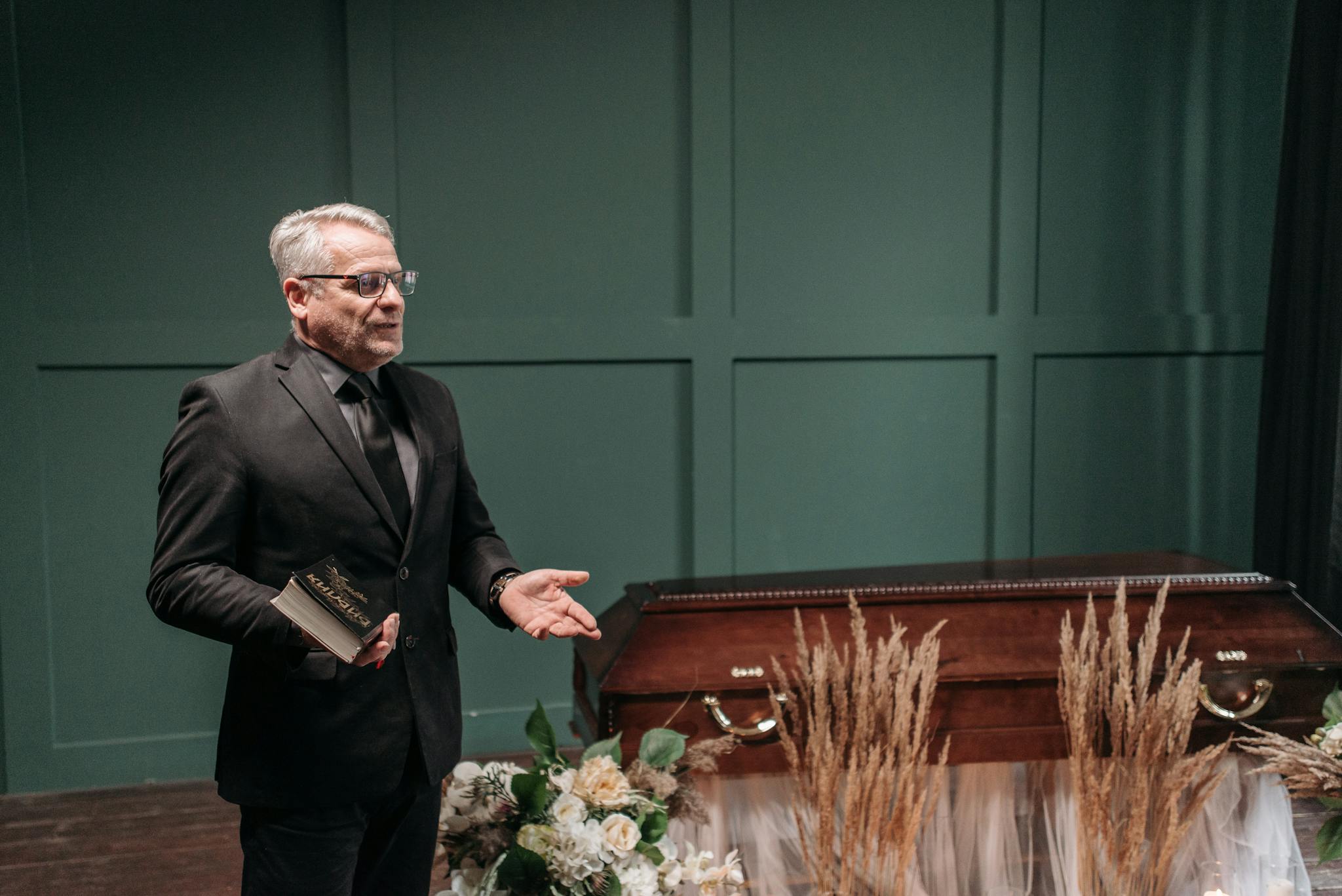 A solemn funeral ceremony featuring a pastor in a black suit delivering a speech next to a wooden coffin adorned with flowers.