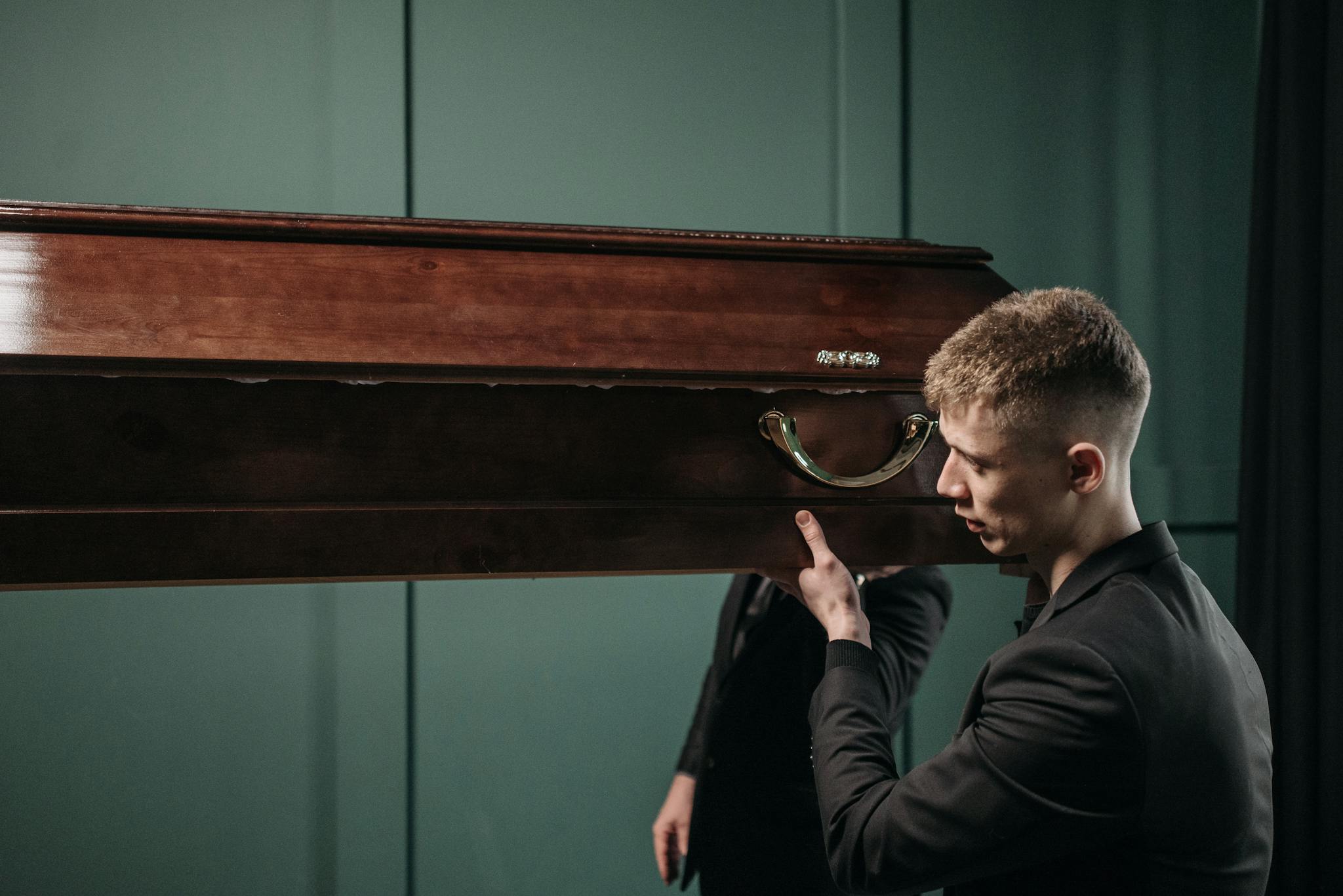 A young man in formal attire carrying a wooden coffin indoors, reflecting a solemn moment.