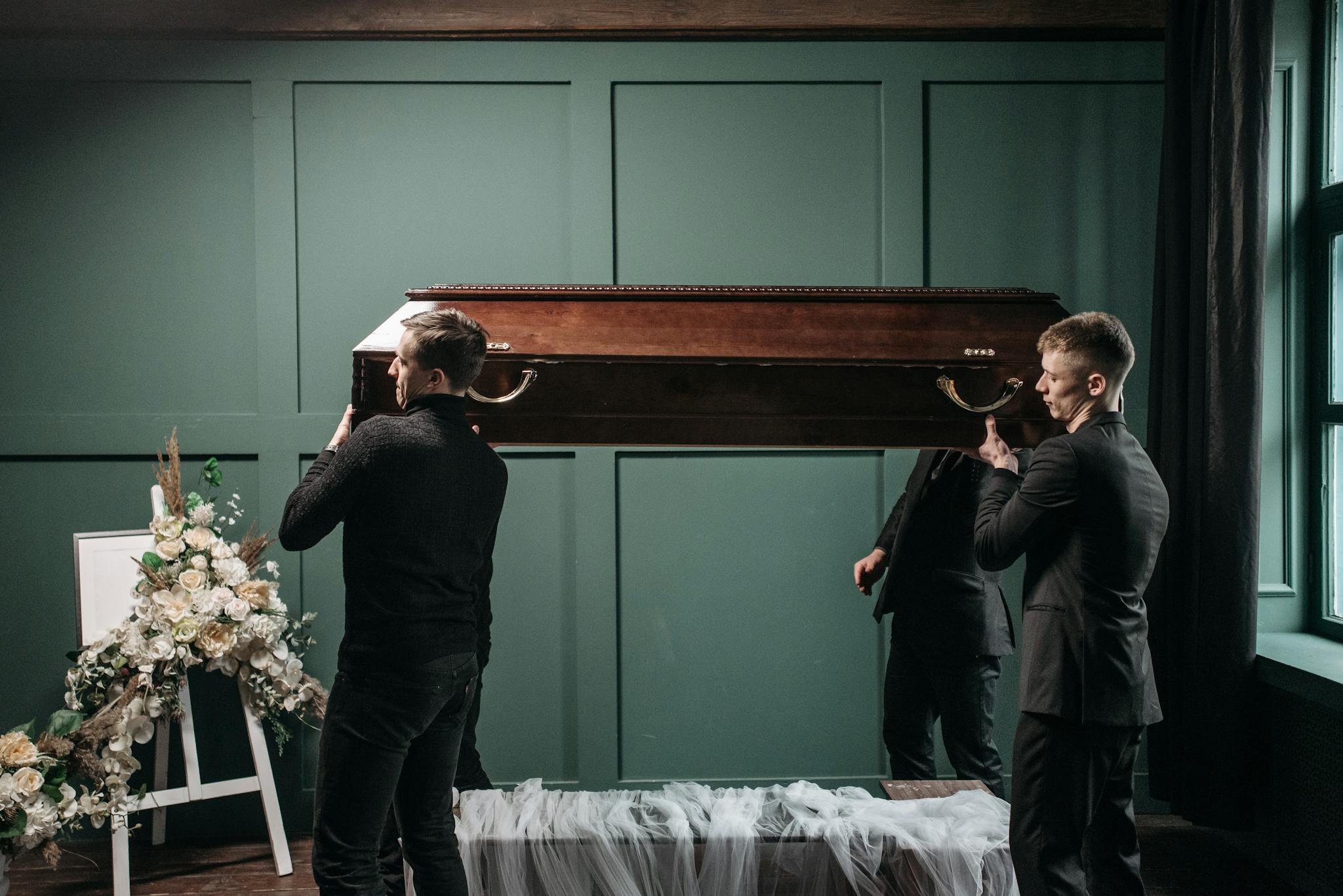 Pallbearers carry a wooden coffin at an indoors funeral service, surrounded by floral tributes.