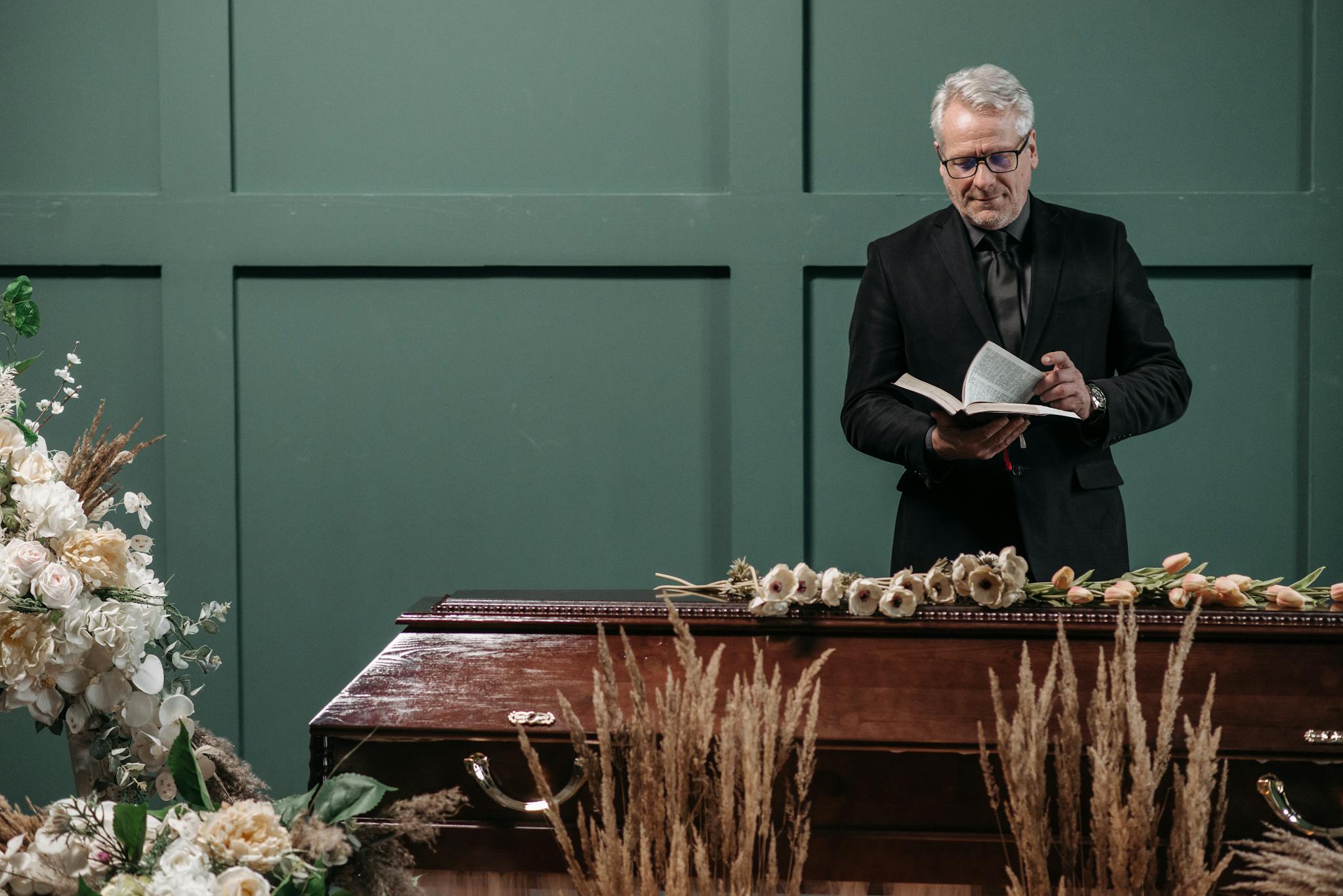 Pastor conducting a funeral service, standing beside a coffin adorned with flowers. Indoor setting.